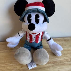 Pu&ograve; includere: Un peluche di Topolino con una camicia a righe rosse e bianche, pantaloncini blu e un cappello con dettagli rossi e blu. Il giocattolo ha orecchie nere, guanti bianchi e piedi grigi. Un'etichetta &egrave; attaccata.