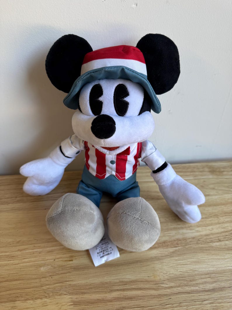 Pu&ograve; includere: Un peluche di Topolino con una camicia a righe rosse e bianche, pantaloncini blu e un cappello con dettagli rossi e blu. Il giocattolo ha orecchie nere, guanti bianchi e piedi grigi. Un'etichetta &egrave; attaccata.
