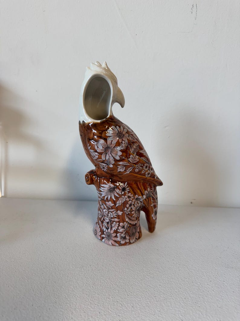 Puede incluir: Un jarr&oacute;n de cer&aacute;mica con forma de loro con base marr&oacute;n y estampado floral blanco. La cabeza del loro es blanca y el jarr&oacute;n tiene una gran abertura. El jarr&oacute;n est&aacute; sobre una superficie blanca.
