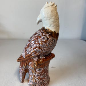 Puede incluir: Figura de loro de cer&aacute;mica posada en un toc&oacute;n de &aacute;rbol. El ave tiene la cabeza y el pico blancos, con patrones florales marrones y blancos en el cuerpo. La base es marr&oacute;n con detalles florales.