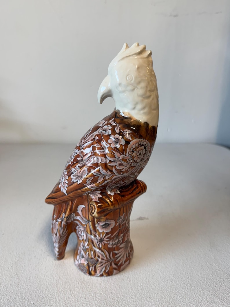 Puede incluir: Figura de loro de cer&aacute;mica posada en un toc&oacute;n de &aacute;rbol. El ave tiene la cabeza y el pico blancos, con patrones florales marrones y blancos en el cuerpo. La base es marr&oacute;n con detalles florales.