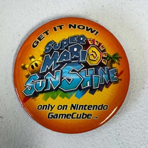 May include: A round orange pinback button featuring the Super Mario Sunshine logo in blue and yellow text. The design includes a small yellow sun icon, a palm tree, and the text GET IT NOW! only on Nintendo GameCube in black lettering.