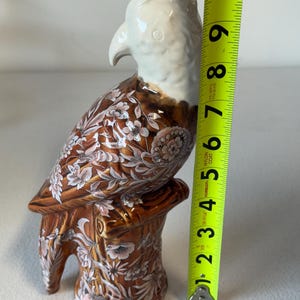 Puede incluir: Una figura de loro de cer&aacute;mica, de aproximadamente 25 cm de altura, posada sobre un toc&oacute;n de &aacute;rbol. El ave presenta una cabeza y un pico blancos, con un cuerpo marr&oacute;n adornado con patrones florales en blanco y rosa. La base tambi&eacute;n es marr&oacute;n con dise&ntilde;os florales.