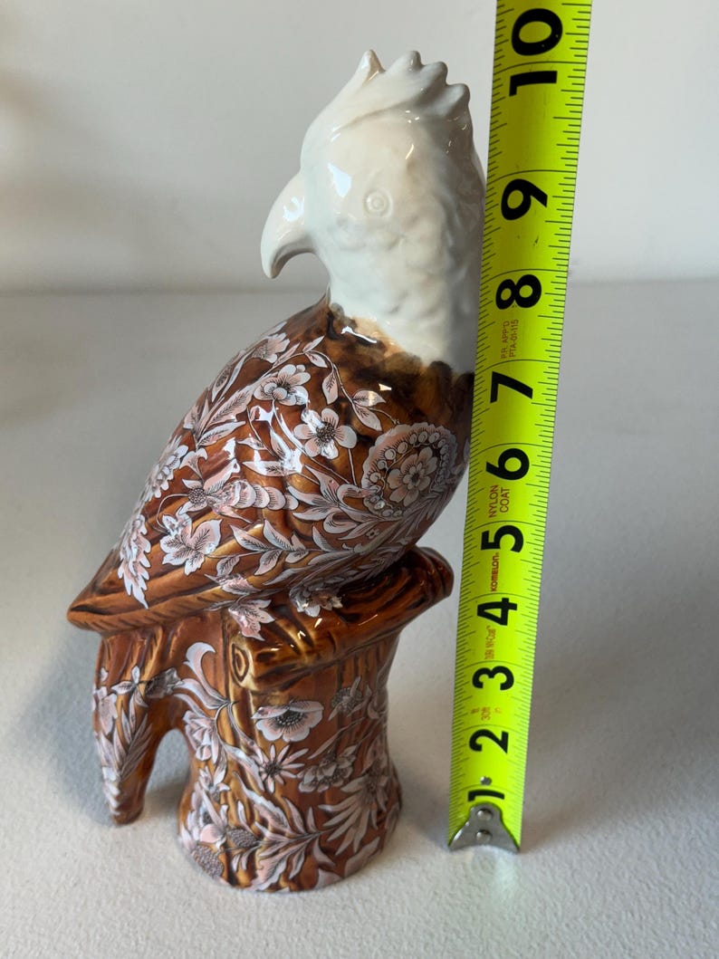 Puede incluir: Una figura de loro de cer&aacute;mica, de aproximadamente 25 cm de altura, posada sobre un toc&oacute;n de &aacute;rbol. El ave presenta una cabeza y un pico blancos, con un cuerpo marr&oacute;n adornado con patrones florales en blanco y rosa. La base tambi&eacute;n es marr&oacute;n con dise&ntilde;os florales.