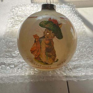 May include: A round, cream-colored glass Christmas ornament featuring a printed illustration of a rabbit wearing a green hat and carrying a bag. The ornament has a silver-toned metal cap and the text Christmas 1979 printed in brown script.
