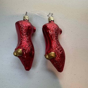 May include: Two red glittery high-heeled shoe ornaments with gold-colored accents at the toe. Each ornament features a metallic cap at the top with a star-shaped loop for hanging, connected by a thin clear string for display on a holiday tree.