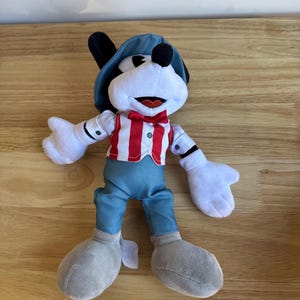 Pu&ograve; includere: Un peluche di un personaggio dei cartoni animati con un viso bianco, orecchie nere e un cappello blu. Il personaggio indossa un gilet a righe rosse e bianche, pantaloni blu e scarpe grigie. Il giocattolo ha guanti bianchi e un papillon rosso.
