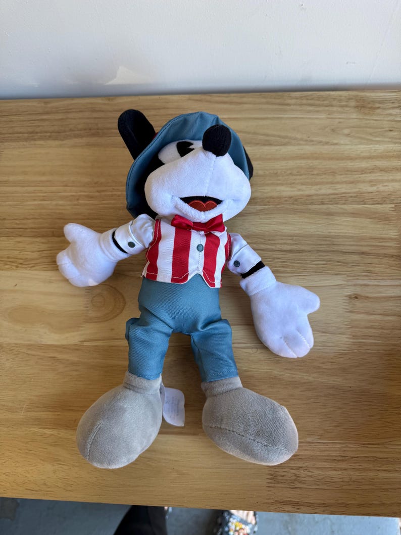 Pu&ograve; includere: Un peluche di un personaggio dei cartoni animati con un viso bianco, orecchie nere e un cappello blu. Il personaggio indossa un gilet a righe rosse e bianche, pantaloni blu e scarpe grigie. Il giocattolo ha guanti bianchi e un papillon rosso.