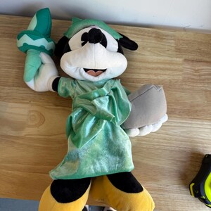 May include: Plush Mickey Mouse dressed as the Statue of Liberty. The plush figure wears a green robe and a matching crown. Mickey holds a torch in his right hand and a tablet in his left. His yellow shoes and black shorts are visible.