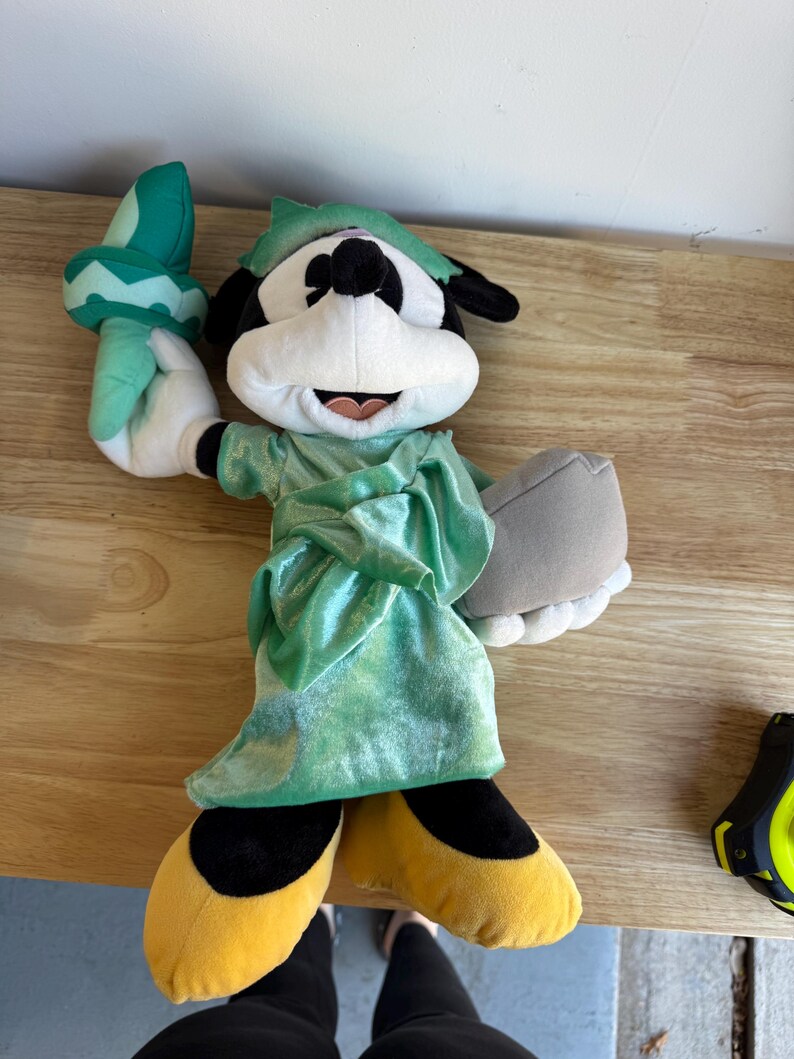 May include: Plush Mickey Mouse dressed as the Statue of Liberty. The plush figure wears a green robe and a matching crown. Mickey holds a torch in his right hand and a tablet in his left. His yellow shoes and black shorts are visible.