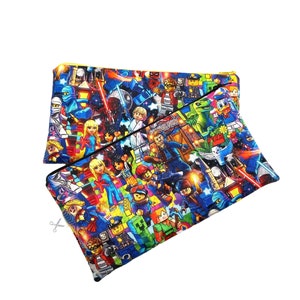 Puede incluir: Una bolsa negra con cremallera con un estampado colorido de varias minifiguras de Lego, incluyendo superhéroes, astronautas y otros personajes. La impresión es sobre un fondo negro con detalles amarillos.