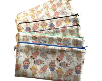Könnte beinhalten: Ein Stapel von fünf rechteckigen Reißverschlusstaschen. Jede Tasche hat ein sich wiederholendes Muster von Cartoon-Hasen in verschiedenen Outfits. Die Taschen haben unterschiedlich farbige Reißverschlüsse: rosa, gelb, schwarz, mintgrün und blau.