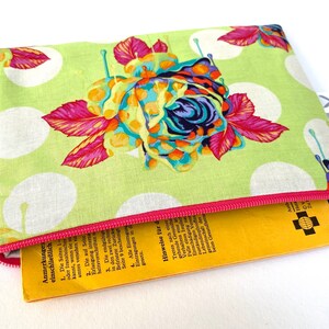 May include: A green zippered pouch with a white polka dot pattern and a colorful floral design. The pouch is partially open, revealing a yellow booklet with German text.