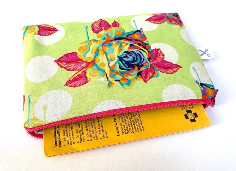 May include: A green zippered pouch with a white polka dot pattern and a colorful floral design. The pouch is partially open, revealing a yellow booklet with German text.