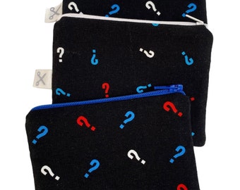 Könnte beinhalten: Drei schwarze Reißverschlusstaschen mit einem sich wiederholenden Muster aus roten, weißen und blauen Fragezeichen. Die Taschen haben unterschiedlich farbige Reißverschlüsse: rot, weiß und blau. Jede Tasche hat ein kleines weißes Etikett mit einer Schere.