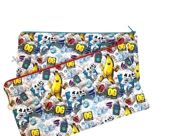 Könnte beinhalten: Zwei Reißverschlusstaschen mit einem sich wiederholenden Muster aus Cartoon-Figuren, Bananen und dem Wort "OG" auf weißem Hintergrund. Die Reißverschlüsse sind blau und rot. Geeignet für kleine Gegenstände.