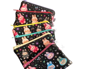 Könnte beinhalten: Eine Reihe schwarzer Reißverschlusstaschen mit einem sich wiederholenden Muster aus Cartoon-Hasen, Regenbogen, Sternen und Blumen. Die Taschen haben unterschiedlich farbige Reißverschlüsse, darunter rosa, gelb und lila. Die Taschen sind aus Stoff.