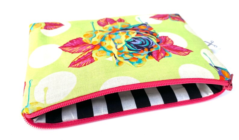 May include: A green zippered pouch with a white polka dot pattern and a colorful floral design. The pouch is lined with black and white stripes.