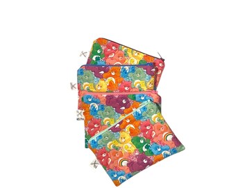 Könnte beinhalten: Mehrere bunte Reißverschlusstaschen mit einem sich wiederholenden Muster von Cartoon-Bären in verschiedenen Farben. Die Taschen haben eine gesteppte Textur und silberne Reißverschlussanhänger. Der Hintergrund ist weiß.
