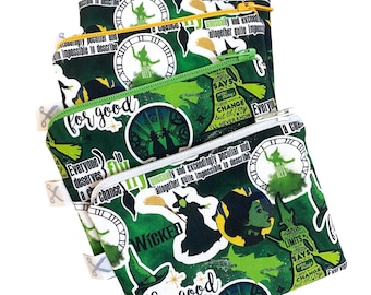 Könnte beinhalten: Drei grüne Reißverschlusstaschen mit einem sich wiederholenden Muster aus Bildern und Texten aus dem Musical "Wicked". Die Taschen zeigen Zitate, Hexensilhouetten und Zifferblätter. Der Text "for good" ist auf einer Tasche zu sehen.