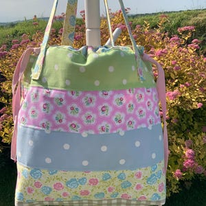 May include: A drawstring tote bag featuring horizontal patchwork panels of light green with white polka dots, pink with floral patterns, light blue with white polka dots, and a bottom band of beige and white gingham, with matching floral straps.