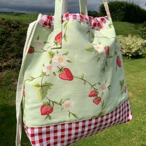 May include: A tote bag featuring a light green fabric with a pattern of red strawberries and white flowers. The bag has a red and white gingham check fabric at the base and a matching red and white gingham drawstring closure at the top.