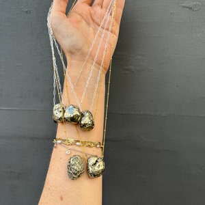May include: A set of five gold necklaces with pyrite stone pendants. The necklaces are layered and feature different chain styles, including a delicate chain and a thicker chain. The pyrite stones are a natural, rough, and irregular shape.