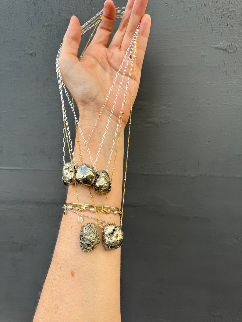 May include: A set of five gold necklaces with pyrite stone pendants. The necklaces are layered and feature different chain styles, including a delicate chain and a thicker chain. The pyrite stones are a natural, rough, and irregular shape.