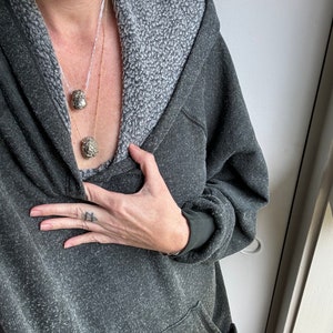 May include: A person wearing a dark gray hooded sweatshirt with a light gray fleece lining. They are wearing a gold chain necklace with two small, round, metallic pendants.