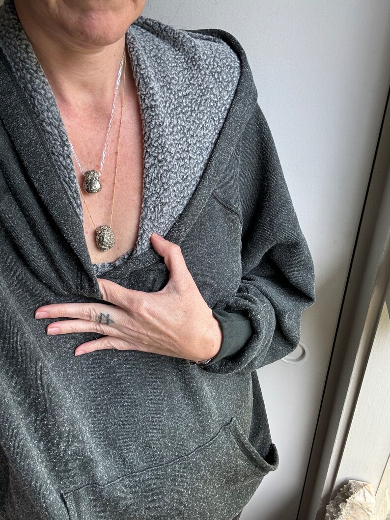 May include: A person wearing a dark gray hooded sweatshirt with a light gray fleece lining. They are wearing a gold chain necklace with two small, round, metallic pendants.