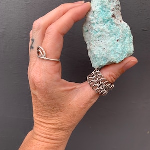 May include: A rough, light green and white rock with brown flecks. The rock is being held in a hand wearing a silver ring and bracelet.