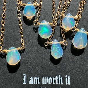 May include: A set of six delicate gold necklaces with iridescent opal teardrop pendants. Each necklace features a simple gold chain and a small opal stone. The necklaces are displayed on a black background with the text "I am worth it" and "www.aliciacoral.com".