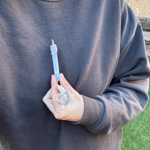 Pu&ograve; includere: Un ciondolo d'argento con un cristallo blu chiaro, avvolto in filo d'argento. Il ciondolo &egrave; tenuto da una mano.