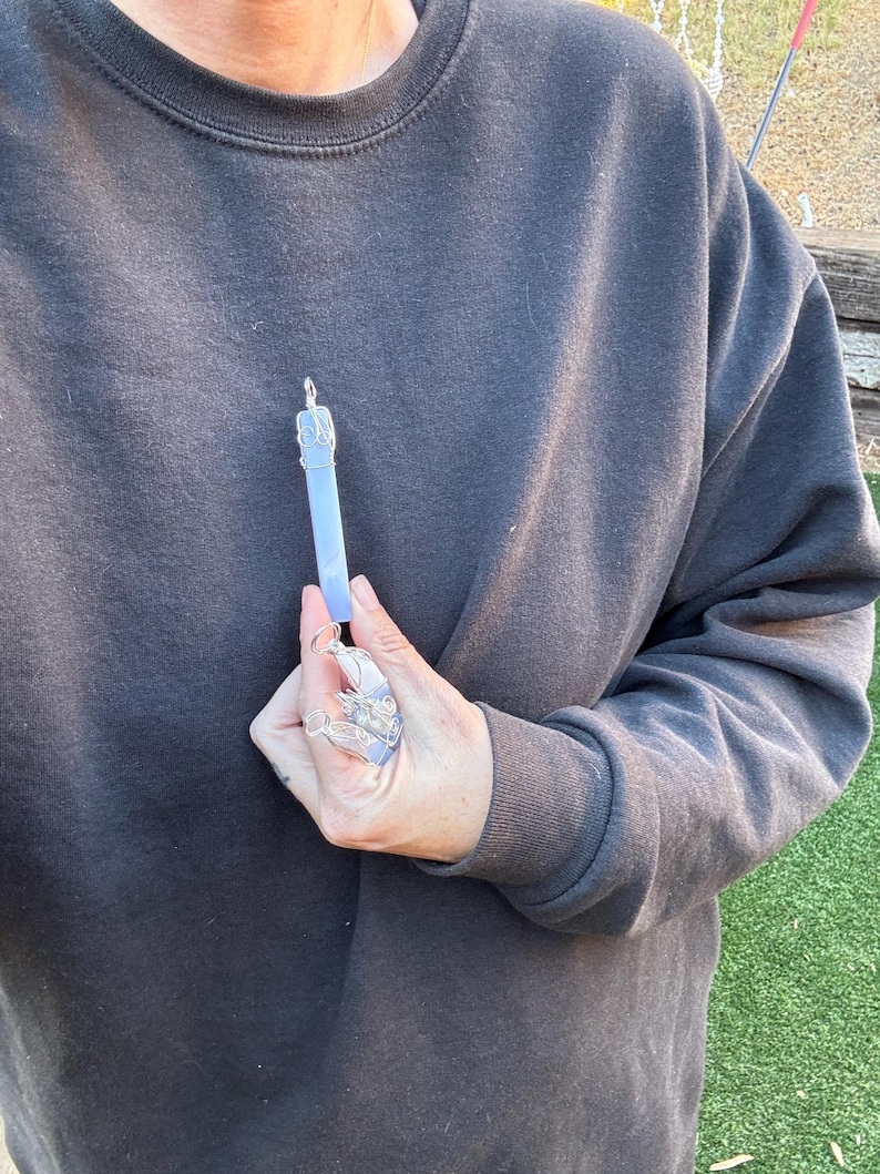 Pu&ograve; includere: Un ciondolo d'argento con un cristallo blu chiaro, avvolto in filo d'argento. Il ciondolo &egrave; tenuto da una mano.