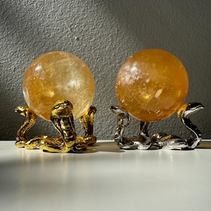 Może przedstawiać: Dwie złote, żółte kryształowe kule na ozdobnych stojakach w kształcie węża. Jeden stojak jest złoty, a drugi srebrny.