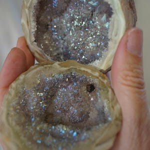 May include: A split geode rock featuring an interior cavity lined with iridescent, light-reflecting crystals. The outer shell of the stone is a smooth, pale beige colour, while the inner crystalline structure displays a shimmering, multi-coloured finish.