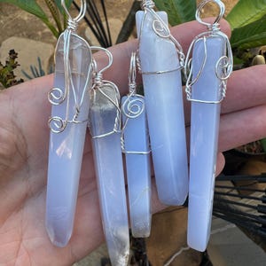 Pu&ograve; includere: Cinque ciondoli in cristallo di calcedonio blu avvolti in filo d'argento. I ciondoli sono di forme e dimensioni diverse e sono appesi a una mano.