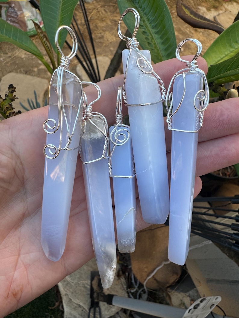 Pu&ograve; includere: Cinque ciondoli in cristallo di calcedonio blu avvolti in filo d'argento. I ciondoli sono di forme e dimensioni diverse e sono appesi a una mano.