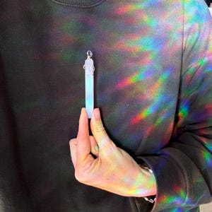 Pu&ograve; includere: Un ciondolo di cristallo bianco con una catena d'argento e una parte superiore decorativa. Il ciondolo &egrave; tenuto da una mano.