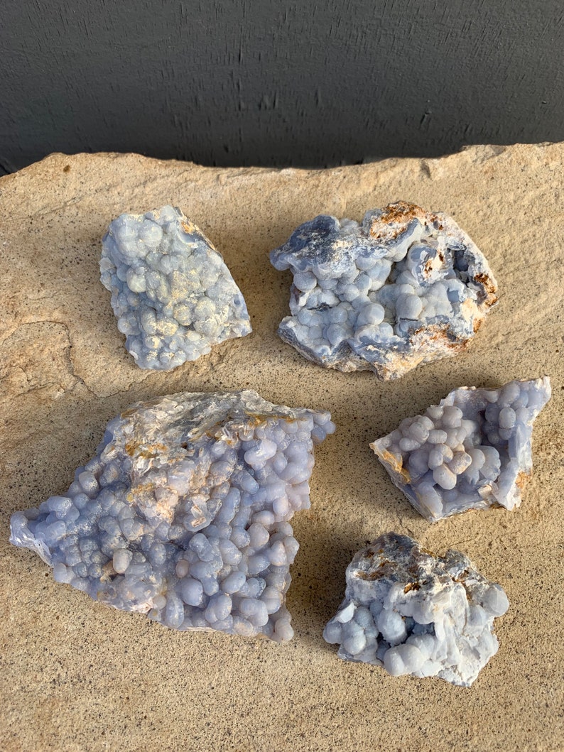 May include: Five blue agate stones with a bubbly texture. The stones are various sizes and shapes and are arranged on a light brown surface.