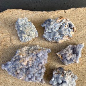 May include: Five blue agate stones with a bubbly texture. The stones are various sizes and shapes and are arranged on a light brown surface.
