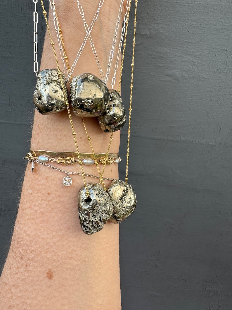 May include: A set of five gold necklaces with pyrite stone pendants. The necklaces are layered and feature different chain styles. The stones are a natural, rough, and textured gray color.