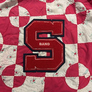 May include: A red and navy blue letter "S" patch with the word "BAND" embroidered on it, sewn onto a pink and white floral quilt. The quilt has a patchwork design with alternating squares and circles.
