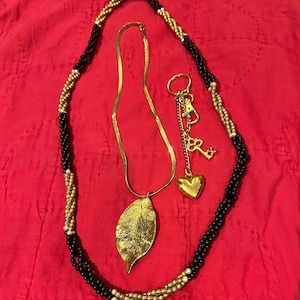 May include: A gold chain necklace with a leaf pendant and a gold heart charm. The necklace is layered with a black and gold beaded chain.