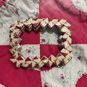 May include: A woven bracelet composed of small, rectangular, folded paper pieces. The paper has a cream background with red diamond shapes and gold accents. The bracelet is displayed on a red and white floral patterned quilt.