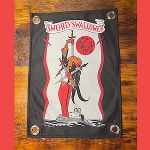May include: A black and white flag with a red and black figure holding swords. The flag has the text "SWORD SWALLOWER" and "Down the Hatch!" in red. The flag has grommets in the corners.