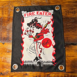 May include: A black and red fabric banner with the text "FIRE EATER" in a white scroll. A red devil with a tail and horns is depicted in a white circle with flames around it. The devil is holding a flaming torch and the text "Hot!" is written in red with a yellow star.