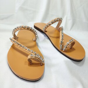 Op de afbeelding: Een paar zandkleurige leren sandalen met meerdere banden versierd met heldere strass steentjes. De sandalen hebben een teenlus en een open achterkant.
