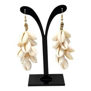 May include: A pair of dangling earrings featuring multiple cream-colored seashells. The earrings are suspended from gold-colored hooks and are displayed on a black stand. The seashells are clustered together, creating a layered effect.