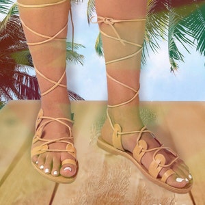 Puede incluir: Sandalias gladiadoras de cuero color canela con cordones que se envuelven alrededor de las piernas. Las sandalias se llevan sobre una superficie de madera con palmeras en el fondo.